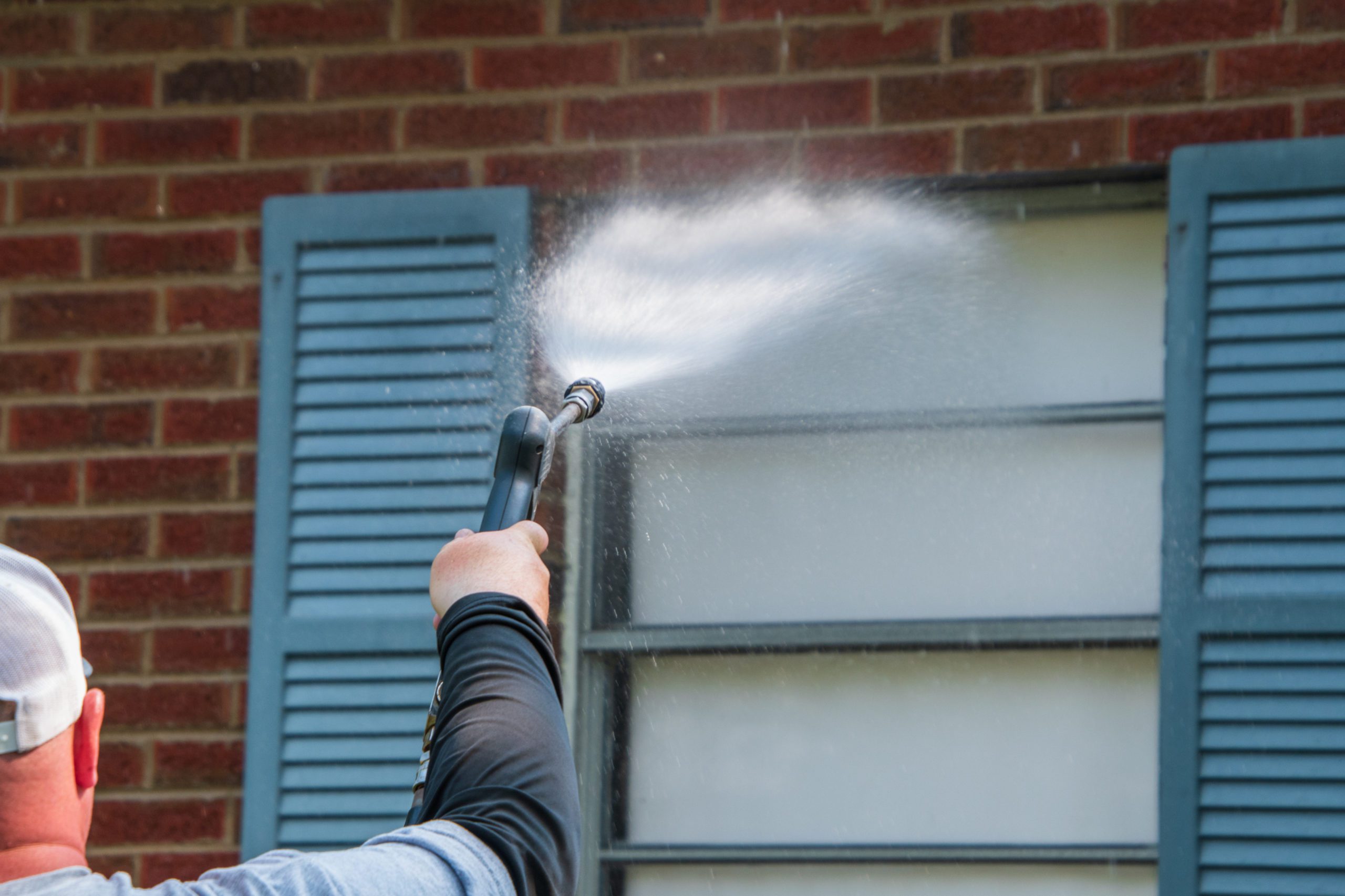 Arm and hand holding a sprayer aimed at a window that has a blue shutter on a red brick wall. The nozzle is spraying a wide water on the window